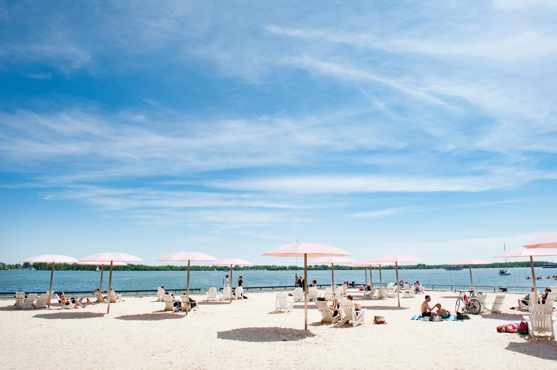 People lounging in chairs under pink umbrellas on the sand at Sugar Beach, Ontario.