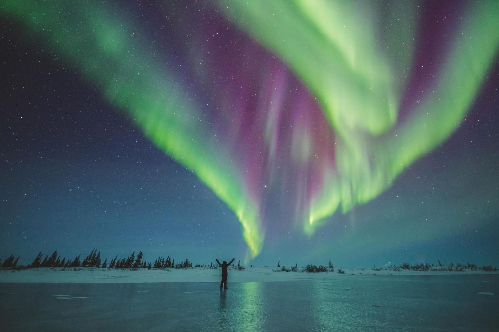 A person stands on a frozen lake, under the northern lights