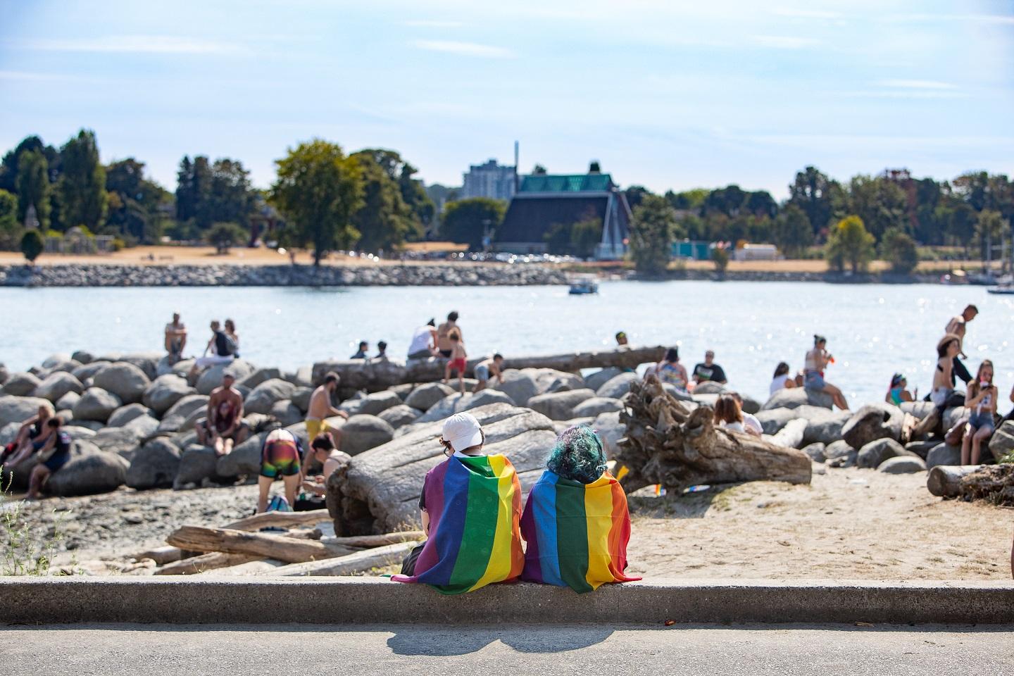Two beachgoers wrapped in pride flags sit on the shore together