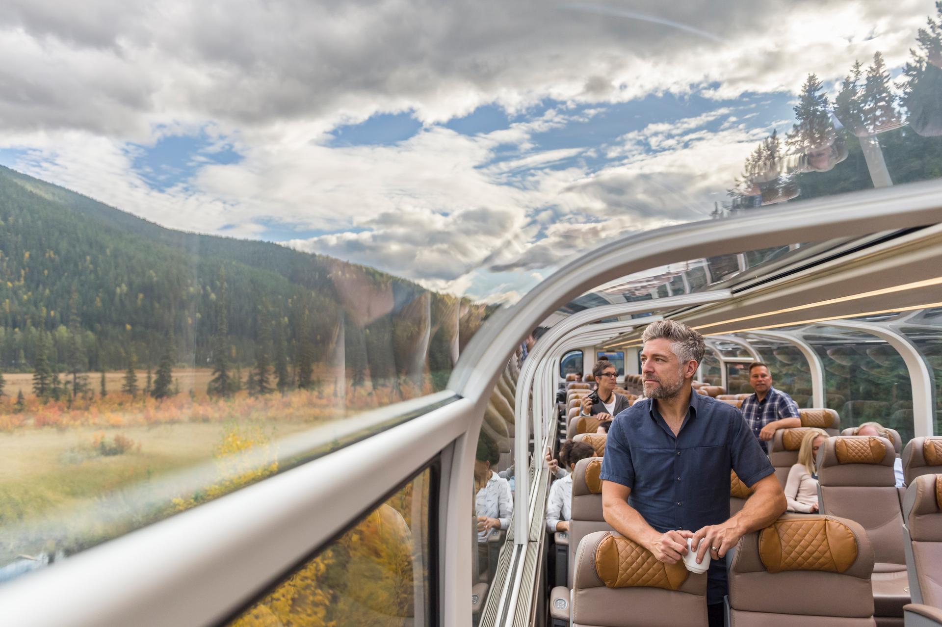 Traveller standing at the panoramic windows of a Rocky Mountaineer Gold Leaf Service glass-dome coach, taking in the forested valleys and mountain scenery of the Canadian Rockies