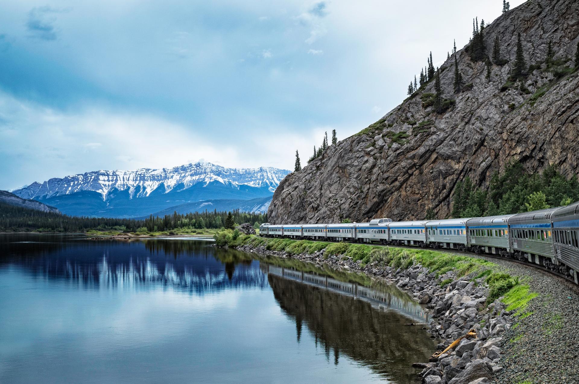VIA Rail passenger train travelling alongside a calm mountain lake reflecting snow-capped Rocky Mountain peaks in the Canadian Rockies