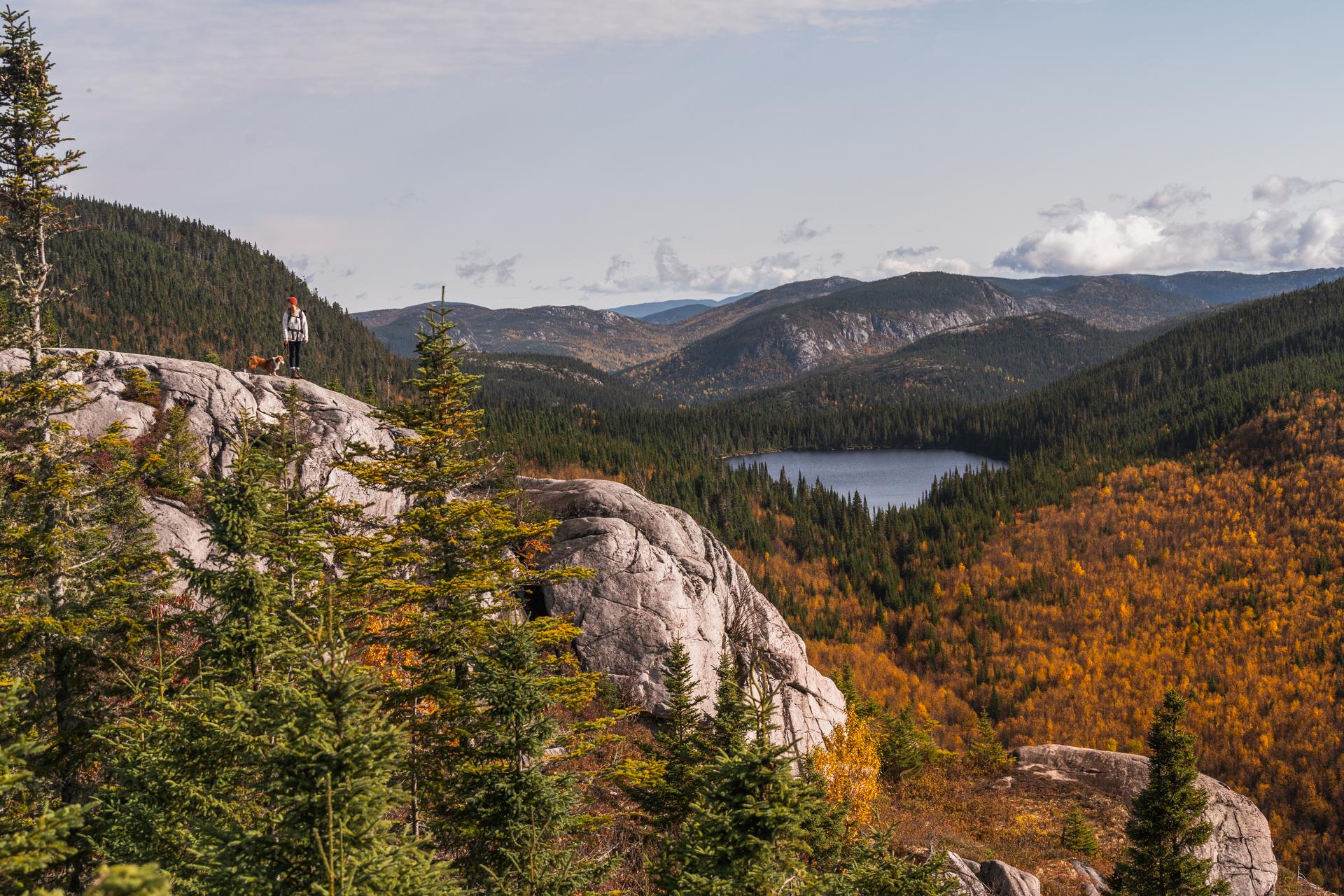 A hiker and dog stand atop a rocky summit overlooking a forest lake and fall foliage along the Charlevoix Traverse in Québec