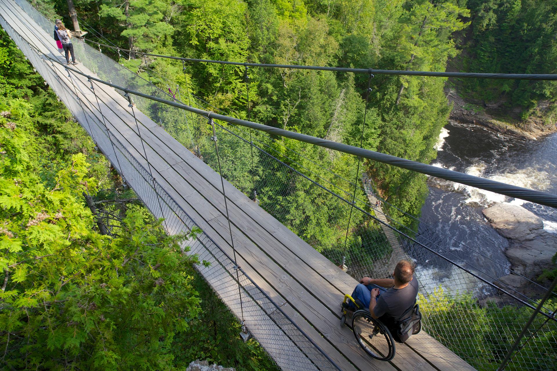 Hikers cross a wooden suspension bridge high above the Sainte-Anne River on the Trans Canada Trail, surrounded by forest